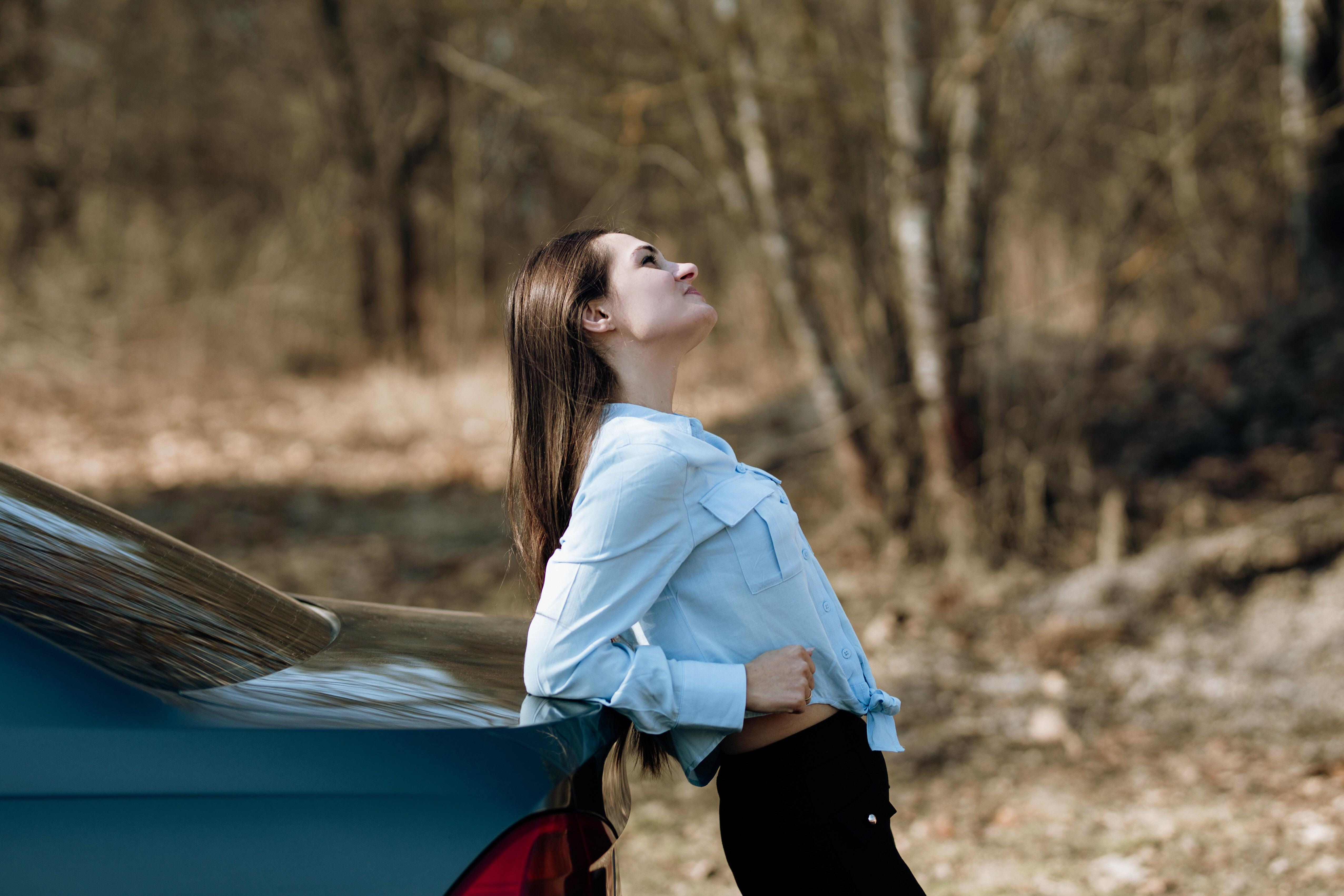 Jak pozować do zdjęć z samochodem? Dziewczyna w jasnej koszuli opiera się o bagażnik samochodu i patrzy w górę - naturalna sesja zdjęciowa z samochodem w lesie